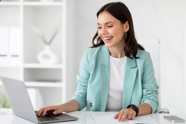 Glad millennial caucasian business lady, manager, secretary in suit typing on laptop at workplace in office interior. Business with device remotely, work and professional occupation