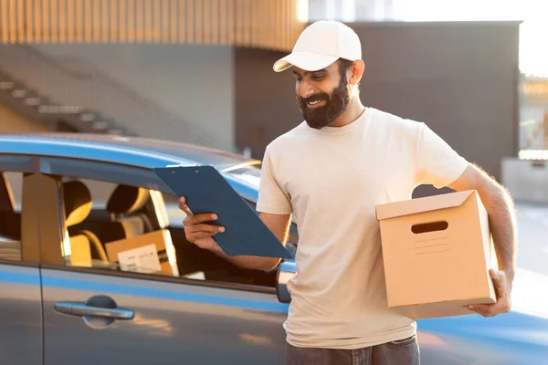 交货报价 快乐的阿拉伯送货员拿着纸板箱 站在外面看剪贴板 头戴帽子 投递客户订单速递服务 — 图库照片