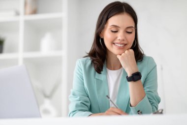 Bin yıllık beyaz takım elbiseli kadın ofisin iç tarafındaki bilgisayarla birlikte iş yerinde notlar alıyor. Aygıt ile iş, yönetici işi, çalışma, webinar ve başlangıç planlaması