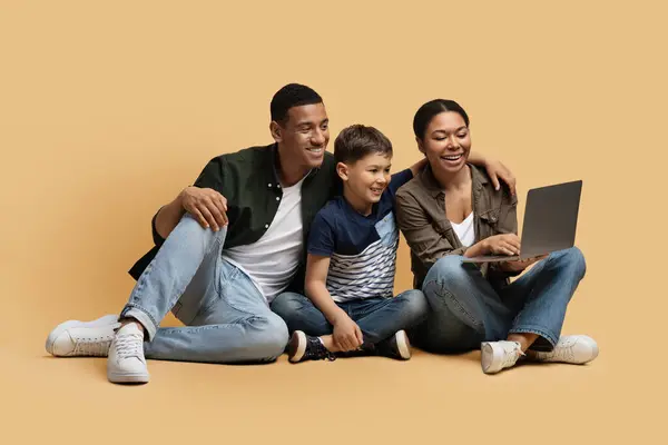 幸せな陽気な若い黒人家族の父親 母親はベージュのスタジオの背景の上に床に座って コンピュータのラップトップを使用してオンライン映画漫画を選択した子供の息子を放棄しました ファミリーエンターテイメント — ストック写真