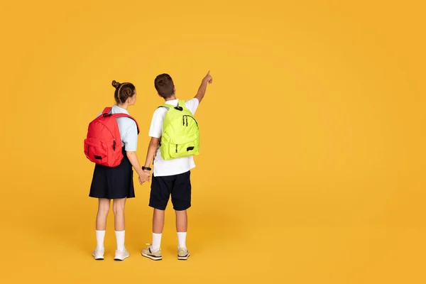 白人少女手牵着男孩儿 手指指向复制空间 隔离在橙色工作室的背景下 好消息 推荐销售 一起学习 学校教育 广告和提供 — 图库照片