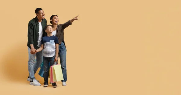 Black friday deal. Happy excited black family millennial parents and school aged boy kid enjoying shopping together, holding colorful purchases and smiling, pointing at copy space, isolated on beige