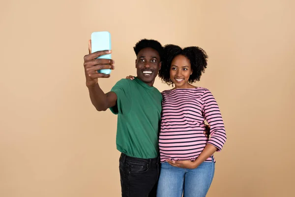 陽気な若い黒人男性は大きな腹で女性を抱擁し ベージュの背景 スタジオで孤立したスマートフォンでセルフィーを作ります 親と家族 ソーシャルメディアアプリ — ストック写真