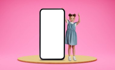 Eğitimli çocuklar için mobil uygulama. Mutlu pozitif okullu küçük kız büyük beyaz ekranlı akıllı telefonun yanında duruyor ve baş parmağını gösteriyor pembe stüdyo arka planında poz veriyor, boşluğu kopyalıyor.