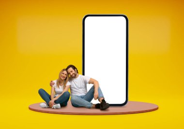 Mutlu neşeli genç çift, sarı stüdyo arka planında, platformda oturuyorlar, beyaz ekranlı büyük telefonun yanında sarılıyorlar, sadece yazlık teklifler sunuyorlar, kopyalama alanı sunuyorlar.
