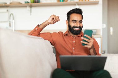 Harika haberlerim var. Neşeli Ortadoğulu adam akıllı telefondan mesaj okuyor dizüstü bilgisayarla oturuyor, evdeki cihazlarla internetten iletişim kuruyor ve çalışıyor. Zafer ve başarı kavramı