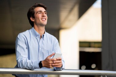 İş uygulaması konsepti. Resmi kıyafetli neşeli genç adam ofis binasının yanında duruyor, elinde cep telefonu, fotokopi odasına bakıyor ve gülümsüyor.