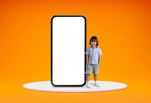 为孩子们提供移动应用程序 可爱而快乐的学龄前小男孩站在讲台上 通过一个巨大的电话 白色的空白屏幕 橙色的工作室背景 空白的空间 — 图库照片