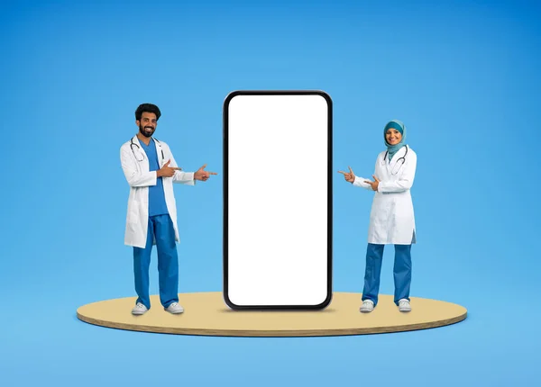 Telemedicine, virtual consultaion, ehealth. Cheerful middle eastern doctors in medical workwear pointing at huge phone with white blank screen, isolated on blue background, mockup, collage
