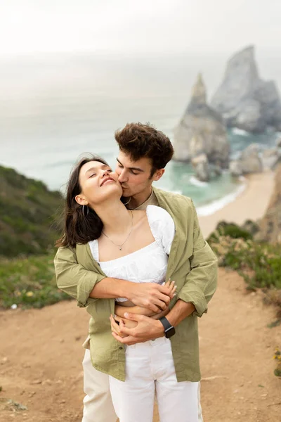 海岸線でデートを楽しむ素敵な若いヨーロッパのカップル 背中から女性を抱擁し 彼女にキス 海岸で完璧な景色で自然に立って垂直ショット — ストック写真