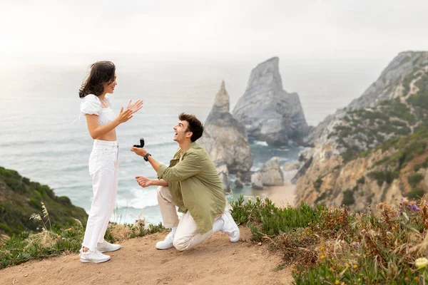 快乐的年轻人向他惊讶的女朋友求婚 跪在地上 欣赏美丽的海岸线 复制空间 户外特别浪漫的婚姻 — 图库照片