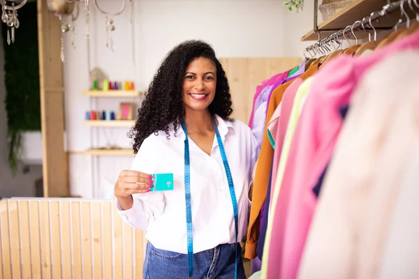 Successful fashion business. Cheerful middle eastern clothing designer lady showing credit card posing with handcrafted garments on rack at dressmaking atelier interior. Commerce and profit concept