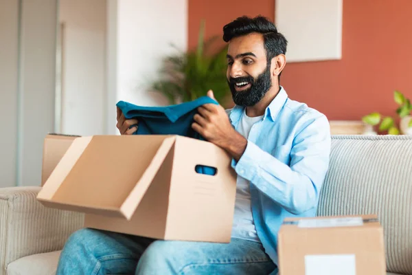 配達サービスとショッピング エキサイティングなアラビアの男は 新しい服を保持し オンラインストアから注文を受信し 自宅のインテリアで配達された段ボール箱を解凍します 選択的なフォーカス — ストック写真