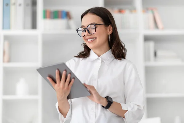 令人高兴的是 年轻的高加索女商人穿着白衬衫 戴着眼镜 在办公室内部的平板电脑上聊天 用于工作的商业和小玩意 官方生活方式 应用程序和广告 — 图库照片