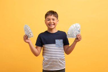 Olumlu, mutlu, okul çağındaki bir çocuk günlük tişört giyiyor elinde nakit para banknotları tutuyor ve sarı arka planda izole bir şekilde gülümsüyor. Çocuk tasarrufu, finansal okur yazarlık kavramı