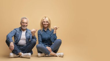 Olumlu neşeli beauitufl yaşlı adam ve kadın sıradan kot elbiseli, bej arka planda izole bir şekilde, fotokopi alanını işaret ederek yerde oturuyorlar. Mutlu evli yaşlı çift reklam yapıyor.