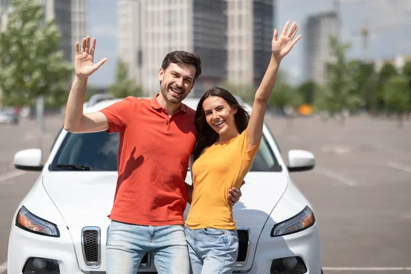 New car, renting, leasing, buying. Thrilled happy young couple wearing casual outfits standing near new white auto, embracing and smiling, raising hands up on street, enjoying new car