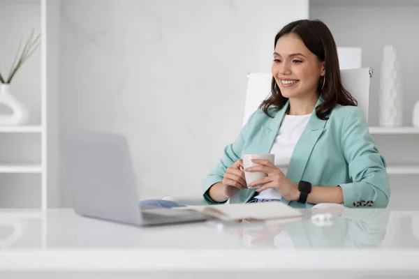 穿着西服的积极的年轻高加索女商人看着电脑 在办公室的内部享受着一杯咖啡 在业务 工作和技术的帮助下 进行远距离会晤 — 图库照片