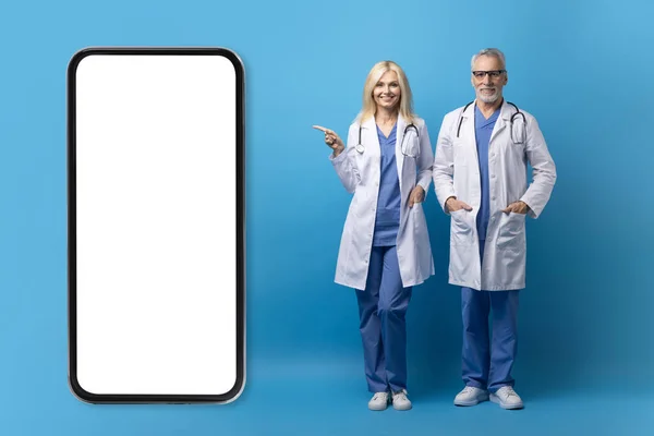 Online appointment with doctor, telehealth concept. Cheerful elderly doctors posing by huge phone with white blank screen over blue studio background, showing healthcare mobile app