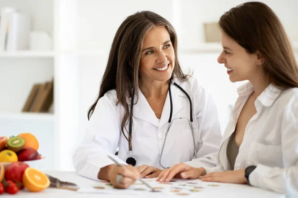 Happy european middle aged lady doctor nutritionist in white coat advises woman patient, recommended diet program in clinic interior. Health care, treatment, weight loss with professional