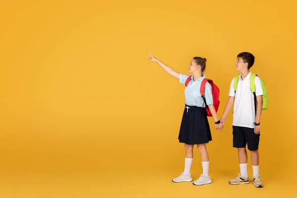 オレンジ色のスタジオの背景に隔離された 空のスペースで指を指す手の少年少年を保持するスマイルな白人少女 推薦研究 学校教育 オファー — ストック写真