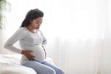 Braxton Hicks contractions. Young black pregnant woman in pain suffering from abdominal ache at home, worried african american female sitting on bed, touching her big tummy, having cramp, copy space clipart