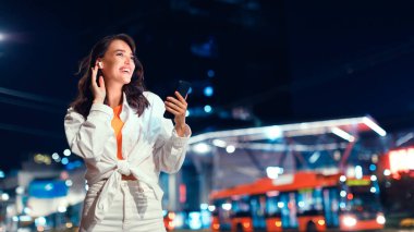 Geceleri şehirde yürürken akıllı telefon kullanan ve kulaklıkla müzik dinleyen genç ve mutlu bir kadın. Kopya alanı olan bir panorama. Metropolün melodileri