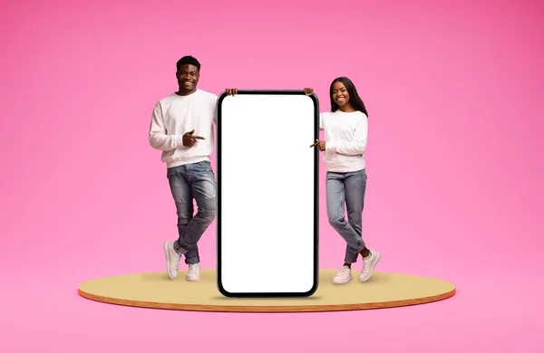 美しい陽気な若いアフリカ系アメリカ人のカップルは ピンクの背景の上にプラットフォーム上にカジュアルに立って 白い空白の画面で巨大な携帯電話を指摘し 素敵なモバイルアプリやオンラインオファーをお勧めします — ストック写真