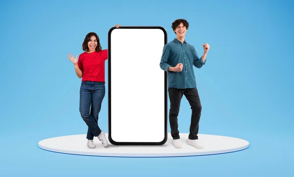 兴奋的中东年轻女人和欧洲女人站在一个大手机旁 手里拿着白色的空白的卷发 广告的空白空间 摆出一副蓝色背景的样子 夫妻们推荐网上提供的好东西 — 图库照片