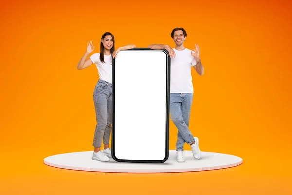 Positive smiling stylish millennial man and woman friends couple standing on platform with huge phone with white blank screen and showing okay, orange background. Great mobile app, mockup