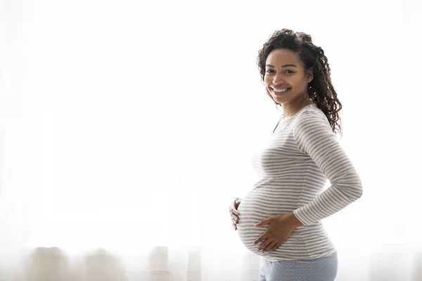 自宅の窓の近くに立っている快適な服で幸せな黒人妊婦 彼女の大きな腫瘍を抱擁するアフリカ系アメリカ人女性に微笑み 赤ちゃんを待って 妊娠時間を楽しんで スペースをコピー — ストック写真