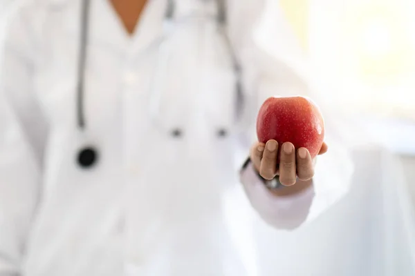 European middle aged lady doctor nutritionist in white coat recommends red apple in clinic interior, close up. Work, professional recommendation and health care, weight loss and diet