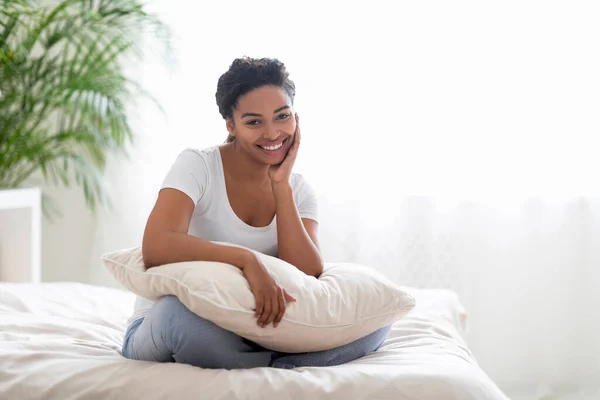 Beautiful young black woman in pajamas sitting on bed, hugging pillow and looking at camera, happy millennial african american female posing in cozy bedroom interior, enjoying home relax, copy space
