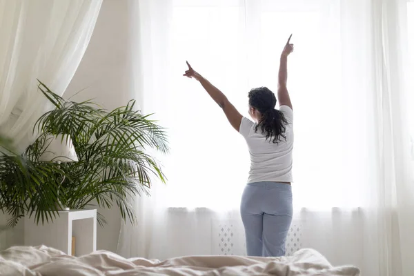 朝起きて家の窓の近くに立っている黒人女性 彼女の体を伸ばすパジャマを身に着けている幸せなエネルギッシュなアフリカ系アメリカ人女性 寝室の快適なベッドの近くでリラックスして 後ろのビュー — ストック写真
