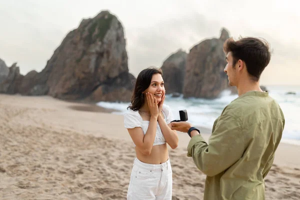 Marry me concept. Young guy making proposal to his surprised girlfriend with ring in gift box, standing on beach during romantic date on coastline, copy space