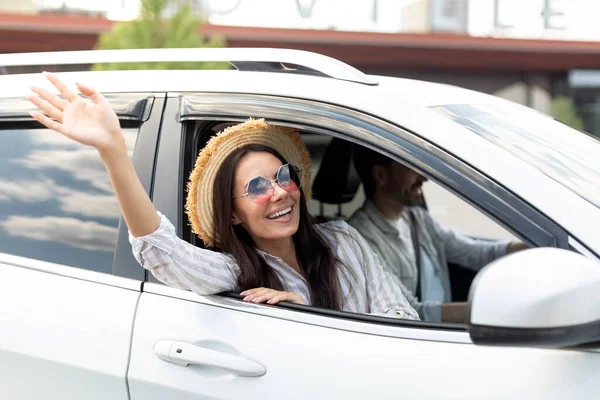かわいいブルネットの若い女性は ウィッカーハットを身に着け 白い車の窓から手を振り 笑顔を浮かべました 陽気な夫婦の配偶者が夏の旅行に行く 一緒にオートバイで休暇 — ストック写真