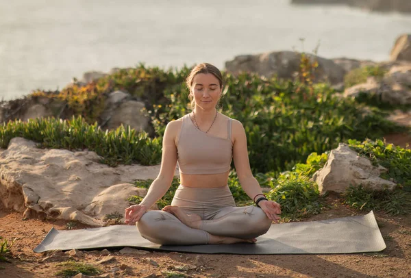 Calm happy millennial caucasian woman athlete in sportswear practices yoga in lotus position, meditates, enjoys workout at sea beach. Sports, body care outdoor and lifestyle at morning