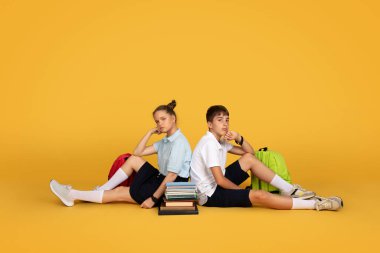 Kafkasyalı üzgün genç kız ve erkek öğrenciler kitaplar, sırt çantaları yerde yatıyor, turuncu stüdyo arka planında izole halde. Okul eğitiminde sorunlar, öğrenim ve mola, reklam, teklif