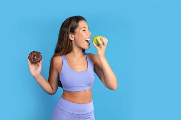 青いスタジオの背景に孤立したドーナツでスポーツウェアの陽気なスリムな若い白人女性 スイーツや適切な栄養 ボディケア ダイエットの選択 — ストック写真