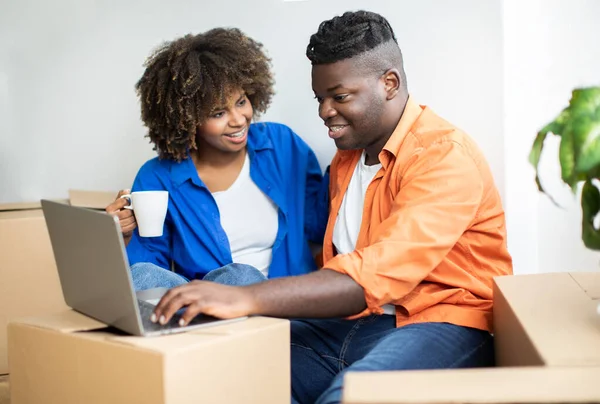 Happy Black Couple Using Computer After Moving To New Home, Smiling African American Spouses Sitting Among Cardboard Boxes In New Flat, Ordering Furniture Online, Purchasing Goods In Internet