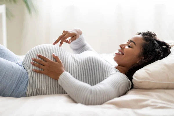 指で歩く黒い妊婦の肖像画は 自宅でベッドに横たわりながら彼女の大きな腹をアクロス 妊娠の時間を楽しんで 妊娠のための準備を期待する女性を微笑みます サイドビュー — ストック写真