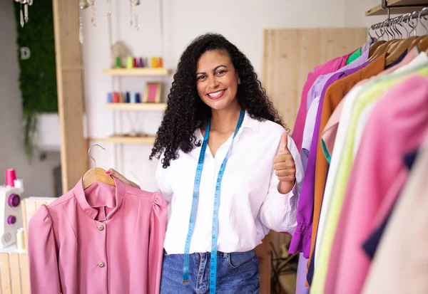I Like Dressmaking. Happy Latin Fashion Designer Lady Showing Her Garment And Thumbs Up Gesture In Clothing Atelier, Standing Near Rack With Clothes. Successful small business, store, commerce