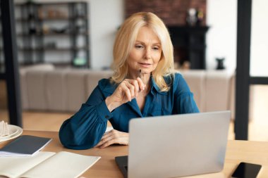 Mesafe çalışması. Evde masa başında dizüstü bilgisayarla çalışan olgun bir serbest çalışan Avrupalı kadın bilgisayar ekranına bakıyor ve düşünüyor.
