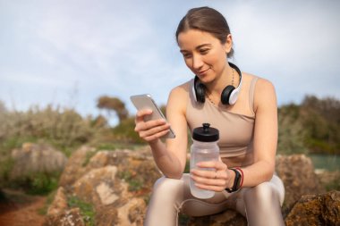 Neşeli bin yıllık beyaz kadın sporcu kadın sporcu, kablosuz kulaklıklar telefonla konuşuyor, taşın üzerinde bir şişe suyla oturuyor. Spor, vücut bakımı uygulaması, sosyal ağlar, blog