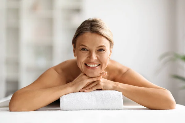 Beautiful middle aged woman lying on table in spa salon, waiting for massage, attractive mature female enjoying wellness treatments and body care, looking and smiling at camera, copy space