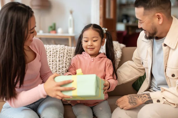 Gifts And Celebration. Asian parents congratulating their little daughter, celebrating birthday holiday and her achievements, giving a special present to child, sitting on sofa at home