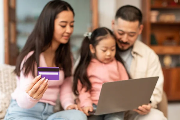 Online Shopping. Happy asian family with child using laptop and credit card, browsing internet stores, ecommerce websites and purchasing items sitting together at home interior. Shallow depth