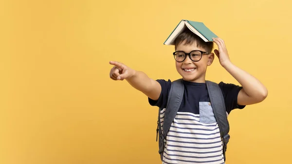 イエローバックグラウンドで隔離されたテキストや広告のコピースペースを指す彼の頭とバックパックの本で眼鏡を身に着けているエキサイティングで陽気なポジティブな少年 パノラマ — ストック写真