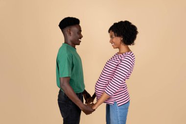 Kocaman göbekli, neşeli, genç siyahi bir adam. Hamilelikten zevk alıyor ve bebek bekliyor. Bej arka planda, stüdyoda izole edilmiş. Ebeveynler ve aile, aşk ilişkisi ve destek