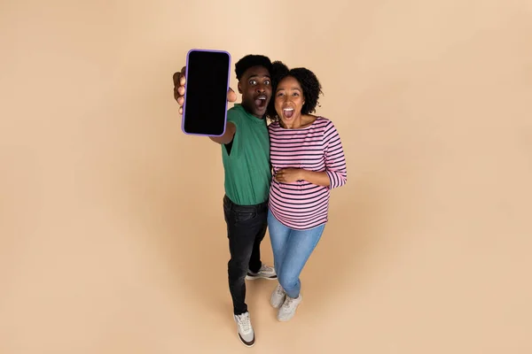 Cheerful young black man hug lady with big belly, shows smartphone with empty screen, isolated on beige background, studio. Parents, family, relationships and pregnancy website, app recommendation